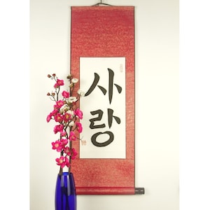 Op de afbeelding: Een rode rol met zwarte kalligrafie die "사랑" leest, wat Koreaans is voor "liefde". De rol hangt aan een witte muur met een vaas met roze bloemen ervoor.