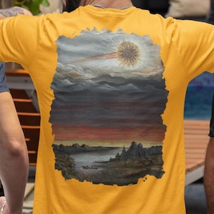 Könnte beinhalten: Drei kurzärmelige T-Shirts in Marineblau, Gelb und Schwarz. Jedes Shirt hat ein Landschaftsdesign auf dem Rücken, das einen Sonnenuntergang über dem Wasser mit einer hellen Sonne und Bergen zeigt. Die Shirts werden von drei Personen getragen.