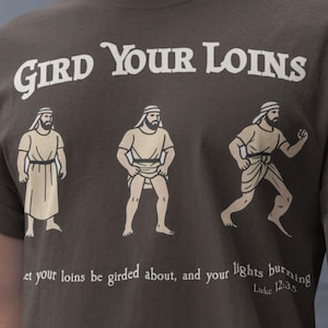 Puede incluir: Camiseta marrón con el texto "GIRD YOUR LOINS" en una fuente vintage. Debajo, tres ilustraciones de hombres con túnicas. La parte inferior de la camiseta tiene texto de Lucas 12:35.