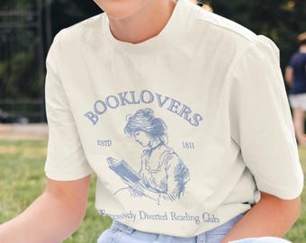 Puede incluir: Una persona con una camiseta color crema con el texto "BOOKLOVERS" y una ilustración de una mujer leyendo. La persona está sentada en una manta sobre la hierba y sosteniendo un libro. La camiseta también tiene el texto "ESTD 1811" y "Exclusively Devoted Reading Club".