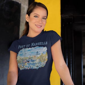 Puede incluir: Camiseta azul marino con la inscripci&oacute;n "Port of Marseille" y una colorida ilustraci&oacute;n de un puerto. La imagen muestra barcos, edificios y una gr&uacute;a, con tonos azules, amarillos y blancos.