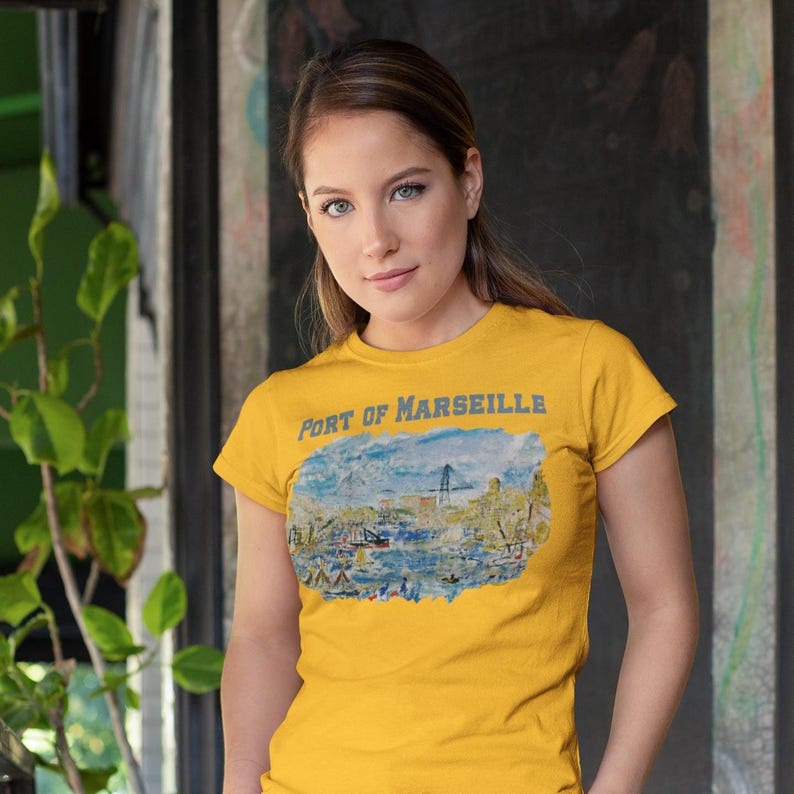 Puede incluir: Camiseta de color amarillo mostaza con el texto "Port of Marseille" sobre una ilustraci&oacute;n de acuarela de una escena portuaria. La obra de arte representa barcos, edificios y un puente. La camiseta tiene mangas cortas.