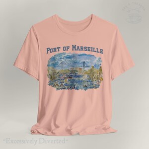 Puede incluir: Camiseta rosa claro con una ilustraci&oacute;n en acuarela del puerto de Marsella. El texto "PORT OF MARSEILLE" est&aacute; impreso en azul encima de la imagen. La frase "Excessively Diverted" est&aacute; impresa en la parte inferior de la camiseta.