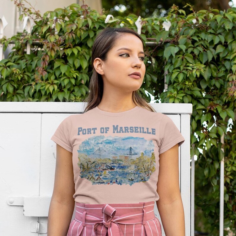 Puede incluir: Una camiseta rosa claro con una ilustraci&oacute;n de acuarela del puerto de Marsella, con el texto "PORT OF MARSEILLE" encima de la imagen. La camiseta la lleva una persona frente a un fondo de hojas verdes.