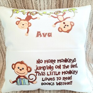 May include: White rectangular pillow with a monkey theme. The top section features two brown monkeys hanging from a green vine, with the name "Ava" below. The bottom section has a monkey reading books and the text "No more monkeys jumping on the bed. This little monkey loves to read books instead!"