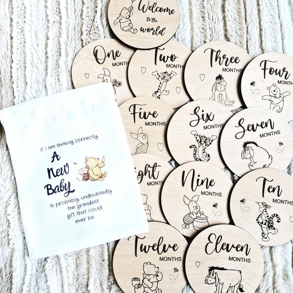 May include: A set of 12 wooden baby milestone discs featuring Winnie the Pooh characters. Each disc has a number from one to twelve, the word "months", and a different character from the Winnie the Pooh franchise. The discs are arranged in a circle on a white fuzzy surface.