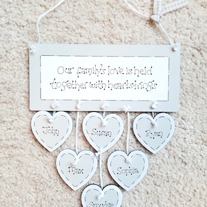 May include: A grey wooden sign with the text "Our family's love is held together with heartstrings". Five white heart-shaped wooden ornaments hang from the sign with the names "John", "Susan", "Ryan", "Max", and "Sophia" written on them. A sixth white heart-shaped ornament hangs below the others with the name "Smudge" written on it.