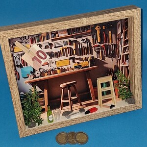 Könnte beinhalten: Schattenbox-Diorama einer Werkstatt mit Werkzeugen an der Wand, einer Werkbank, einem Hocker und einer kleinen Leiter. Ein 10-Euro-Schein ist an der Werkbank befestigt. Eine kleine Flasche, ein Glas und Münzen befinden sich im Vordergrund.