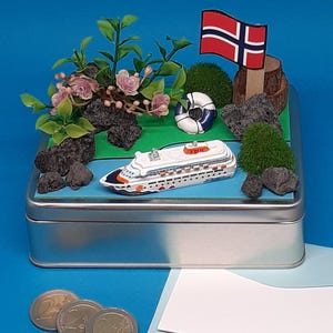 Könnte beinhalten: Eine silberne Metallbox mit einer Miniatur-Szene im Inneren. Die Szene enthält ein kleines Kreuzfahrtschiff, einen Rettungsring, einen kleinen grünen Busch, einen kleinen Holzstamm, eine norwegische Flagge und schwarze Steine. Die Box steht auf einer blauen Oberfläche.