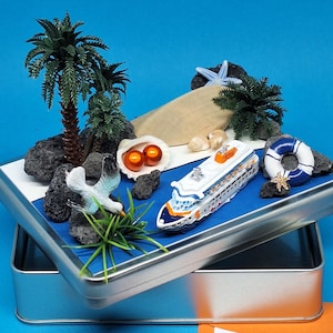 Könnte beinhalten: Eine Miniatur-Strandszene in einer silbernen Blechdose. Die Szene umfasst ein Spielzeug-Kreuzfahrtschiff, Palmen, eine Möwe, eine Rettungsboje und Muscheln. Der Hintergrund ist blau.