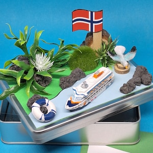 Könnte beinhalten: Miniatur-Diorama in einer silbernen Dose mit einem Kreuzfahrtschiff, einer norwegischen Flagge, einer Möwe und einer Rettungsboje. Die Szene umfasst grünes Laub, Felsen und einen blauen Hintergrund, der ein maritimes Thema erzeugt.