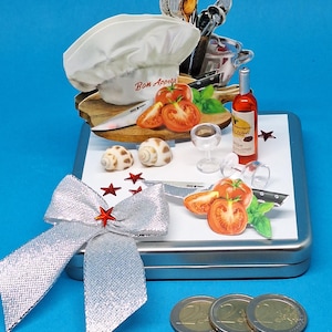 Könnte beinhalten: Eine dekorative Geschenkbox zum Thema Kochen mit einer Kochmütze mit dem Text "Bon Appetit", Tomaten, einer Weinflasche und einer silbernen Schleife. Enthält Miniatur-Küchenutensilien, Weingläser und Sternakzente. Die Box ist ca. 10 cm lang.