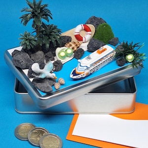 Könnte beinhalten: Eine Miniatur-Strandszene in einer silbernen Dose. Die Szene zeigt ein Spielzeug-Kreuzfahrtschiff, Palmen, eine Möwe, Felsen, Muscheln und eine Rettungsboje. Euro-Münzen und ein orangefarbener Umschlag befinden sich im Vordergrund.