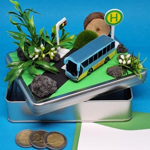 Könnte beinhalten: Miniatur-Diorama in einer silbernen Dose, mit einem blau-gelben Bus, einer Ampel und einem Bushaltestellenschild. Die Szene umfasst Grün, Felsen und ein Straßendesign. Im Vordergrund sind drei Euro-Münzen vor blauem Hintergrund zu sehen.