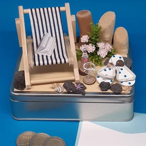 Könnte beinhalten: Eine Miniatur-Strandkulisse in einer silbernen Blechdose. Die Szene beinhaltet einen gestreiften Strandstuhl, ein Buch, ein Glas Wein, Flip-Flops und eine kleine Topfpflanze mit rosa Blüten. Die Dose steht auf einer blauen Oberfläche mit drei Münzen davor.