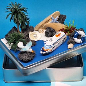 Könnte beinhalten: Ein Miniatur-Kreuzfahrtschiff, Palmen und eine Möwe sind auf einer blauen Oberfläche in einer silbernen Dose angeordnet. Dekorative Elemente sind eine Muschel, Treibholz und eine Rettungsboje. Das Thema ist Urlaub.