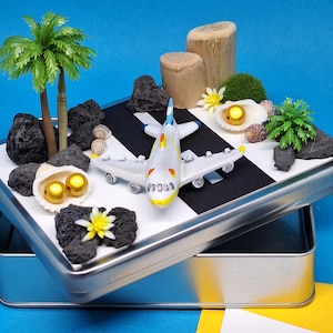 Könnte beinhalten: Ein Miniaturflugzeug auf einer schwarz-weißen Startbahn, eingebettet in einer silbernen Dose. Die Szene umfasst Miniaturpalmen, Felsen, Muscheln und eine kleine Blume. Das Flugzeug ist weiß mit gelben und blauen Akzenten. Die Dose steht auf einer blauen Oberfläche.