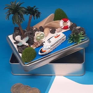 Könnte beinhalten: Eine Miniatur-Strandszene in einer silbernen Blechdose. Die Szene zeigt ein Spielzeug-Kreuzfahrtschiff, Palmen, Muscheln, Felsen und eine Rettungsboje. Zwei Euro-Münzen befinden sich im Vordergrund. Der Hintergrund ist einfarbig blau.