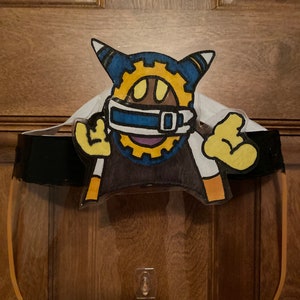 May include: A clear plastic face shield with a yellow border. A cartoon character with a blue, yellow, and white design is attached to the shield.
