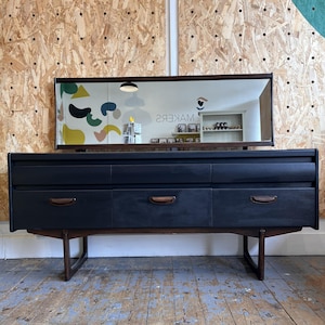 May include: A dark charcoal sideboard featuring six drawers with integrated wooden handles, supported by a wooden frame. A rectangular mirror is attached to the top, reflecting the surrounding space and the text MAKERS in reverse.