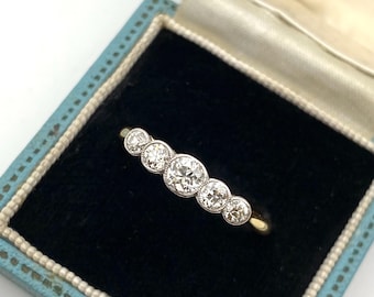 Könnte beinhalten: Ein Ring mit fünf runden Diamanten im Brillantschliff, die in einer Reihe in einer Metallschiene gefasst sind. Der Ring liegt in einer hellblauen, rechteckigen Schmuckschatulle mit schwarzem Samtfutter und weißem Rand an den Kanten.