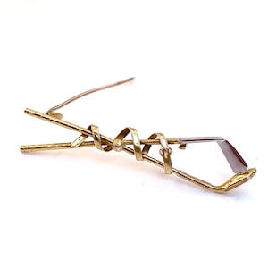 May include: A gold-toned metal brooch in the shape of two golf clubs intertwined. The clubs are twisted together and the ends of the clubs are visible.