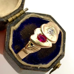 May include: A gold ring with a heart-shaped design, featuring a ruby and a diamond. The ring is displayed in a vintage blue velvet jewellery box. The ring's band and heart shapes are a warm gold colour.