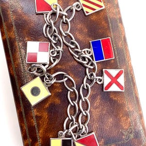 Könnte beinhalten: Ein silbernes Charm-Armband mit nautischen Signalflaggen in Rot, Gelb, Weiß und Blau. Die quadratischen Flaggen sind durch eine Kette verbunden. Das Armband wird auf einer braunen, strukturierten Oberfläche präsentiert.