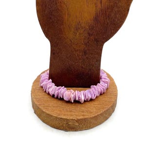 Puede incluir: Un brazalete de concha morado claro exhibido en un expositor de joyas de madera. El brazalete está hecho de piezas de concha irregulares y pulidas en tonos lavanda y rosa. El soporte es de color madera natural y el fondo es blanco.