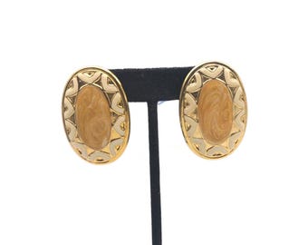 May include: A pair of oval-shaped earrings with a gold-tone frame. The center of each earring features a light brown, swirled design, surrounded by a cream-colored border with a geometric pattern. The earrings are displayed on a black stand.