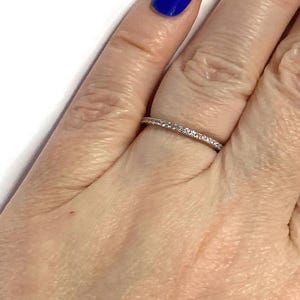 Könnte beinhalten: Ein dünner, silberner Ring mit einer Reihe kleiner, klarer Edelsteine. Der Ring befindet sich an einem Finger mit blauem Nagellack. Ein zarter Schmuck.