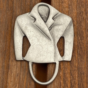 Puede incluir: Un colgador de pared de metal plateado con forma de abrigo. El colgador es una representación estilizada de una chaqueta con cuello y solapas. El colgador está montado sobre una superficie de madera con tinte marrón oscuro.