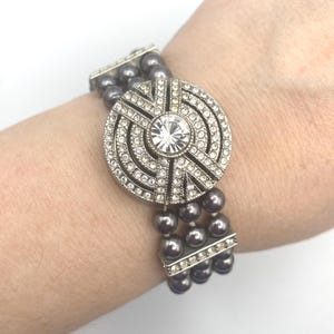 Puede incluir: Una pulsera plateada con tres hilos de cuentas grises. El centro presenta un gran adorno redondo plateado con una piedra central clara y facetada y cristales claros circundantes. La pulsera se lleva en la muñeca.