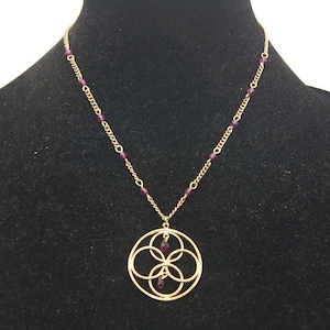 May include: A gold necklace with a pendant featuring a circular design with four overlapping circles and purple gemstones. The necklace is on a black velvet display stand.