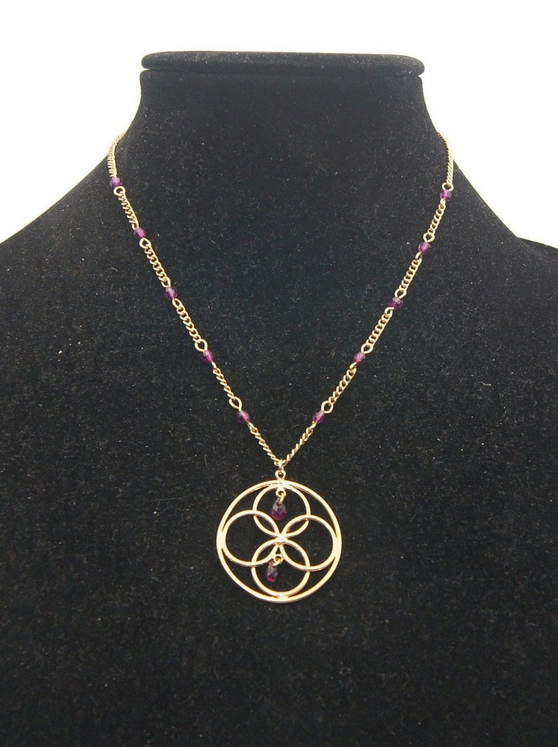 May include: A gold necklace with a pendant featuring a circular design with four overlapping circles and purple gemstones. The necklace is on a black velvet display stand.