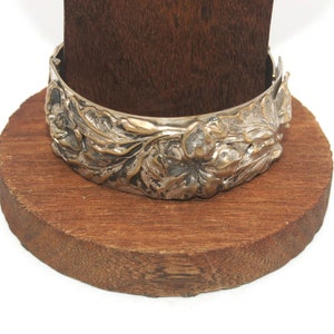Könnte beinhalten: Ein silbernes Armband mit floralem Design. Das Armband befindet sich auf einem Holzständer.