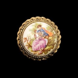 Puede incluir: Un botón redondo de color dorado con un centro de porcelana blanca que presenta una escena pintada de una pareja en un entorno de jardín.