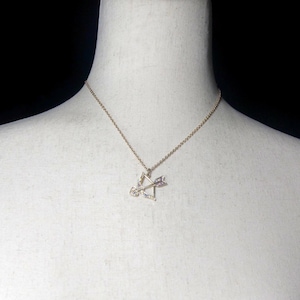 May include: A gold chain necklace with a bow and arrow pendant. The pendant is decorated with small, clear crystals.