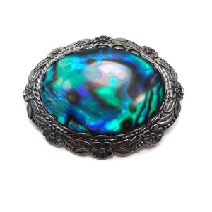 Op de afbeelding: Een zilverkleurige broche met een grote ovale inleg van abalone-schelp. De schelp heeft iriserende blauwe, groene en paarse kleuren. De broche is versierd met een bloemmotief.