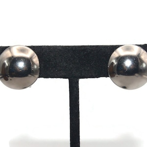 May include: A pair of silver tone round stud earrings. The earrings are on a black display stand.