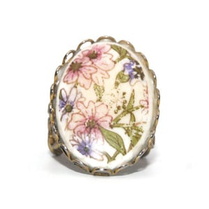 Puede incluir: Un anillo vintage con un camafeo ovalado de cerámica blanca con un diseño floral con flores rosas, moradas y verdes. El camafeo está engastado en un marco de metal dorado con un borde festoneado.