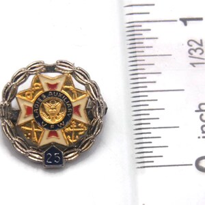 K&ouml;nnte beinhalten: Eine Vintage-Anstecknadel der Ladies Auxiliary VFW. Die Anstecknadel ist rund, mit einem zentralen Emblem mit einem Adler und dem Text "Ladies Auxiliary VFW". Sie hat ein wei&szlig;es, rotes und goldenes Sternmuster, umgeben von einem silbernen Kranz und der Zahl "25".