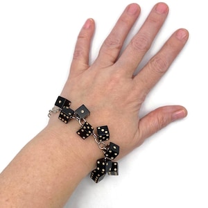 May include: A black dice bracelet with white dots, connected by silver-colored chain links. The bracelet is shown on a person's wrist against a white background. Each die has a different number of dots.