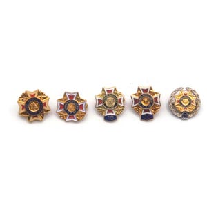 K&ouml;nnte beinhalten: F&uuml;nf verzierte, goldfarbene Broschen mit aufwendigen Designs und farbenfrohen Akzenten. Jede Brosche hat ein zentrales Emblem, das von einem detaillierten Rand umgeben ist, mit roten, blauen und wei&szlig;en Emaille-Details. Die Broschen sind auf wei&szlig;em Hintergrund angeordnet.