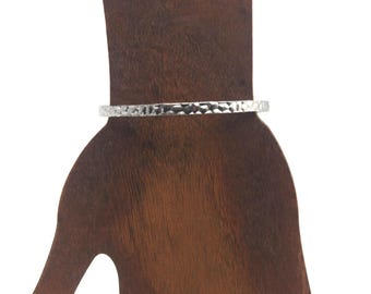 Op de afbeelding: Een zilverkleurige gehamerde metalen armband. De armband wordt getoond op een bruine houten vorm. De armband heeft een textuur en een eenvoudig ontwerp. De sieraden zijn geschikt voor dagelijks gebruik.