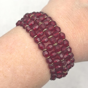 May include: A multi-strand bracelet featuring deep red, faceted, translucent beads. The beads are arranged in four parallel rows, separated by thin, metallic gold-toned wire spacers that connect the individual beads in a structured, repeating pattern.