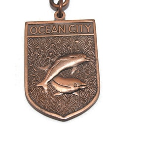 May include: A copper-colored keychain with a shield-shaped charm. The charm features the text "OCEAN CITY" and two dolphins jumping out of the water.