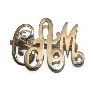 May include: Gold-toned metal monogram hair clip with the letters "AM" in a decorative script font.