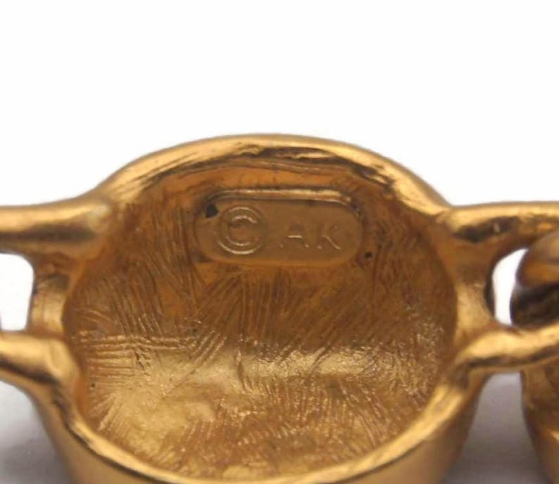 May include: Close-up of a gold-toned metal chain with a round link featuring the inscription "&copy; AK".