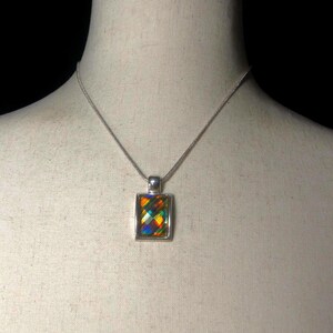May include: A silver necklace with a rectangular pendant. The pendant features a colorful, mosaic-like design with shades of orange, yellow, green, and blue. The necklace is displayed on a mannequin.
